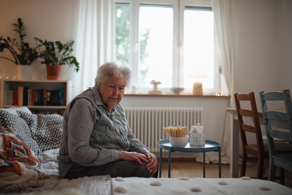 Elderly woman sitting alone on a couch in a quiet living room, looking down thoughtfully