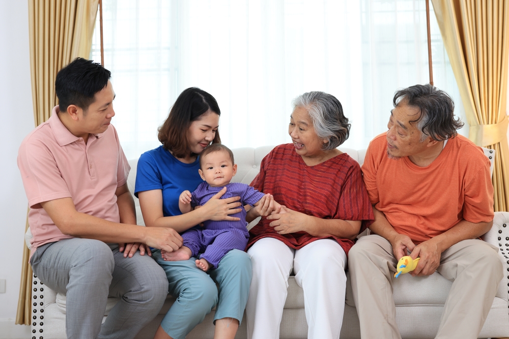 Three generation family — grandparents, parents, and a baby — sitting together on a sofa in a bright living room